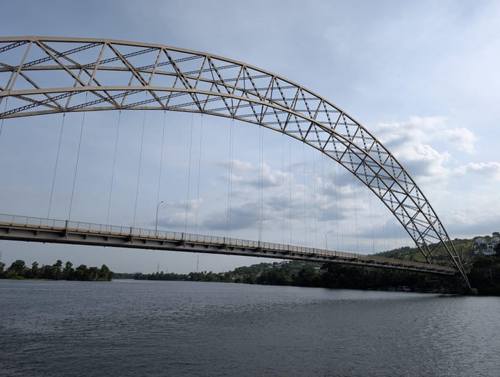Stahlbogenbrücke über einen ruhigen Fluss unter bewölktem Himmel, von unten fotografiert sodass der geschwungene Träger den Bildrahmen dominiert.